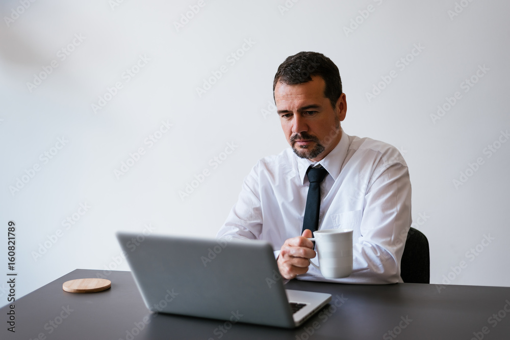 Senior businessman using laptop computer at wor, holding cup of coffee Senior businessman using laptop computer at wor, holding cup of coffee