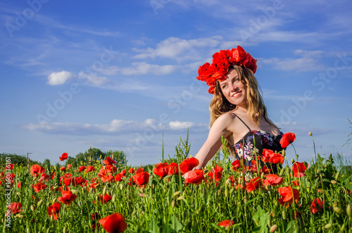 Young charming girl walks on the field with flowering poppies with a wreath of poppies on her head