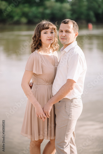 beautiful young couple near a large river, cuddling, standing watching with a smile, pressed against each other, in nature. summer, walk