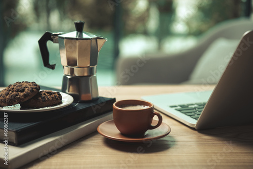 Close-up view of laptop, cup of coffee, moka pot and chocolate chip cookies with books on wooden table photo