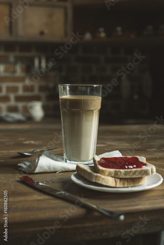 latte in glass, bread with jam on plate and cutlery on wooden tabletop