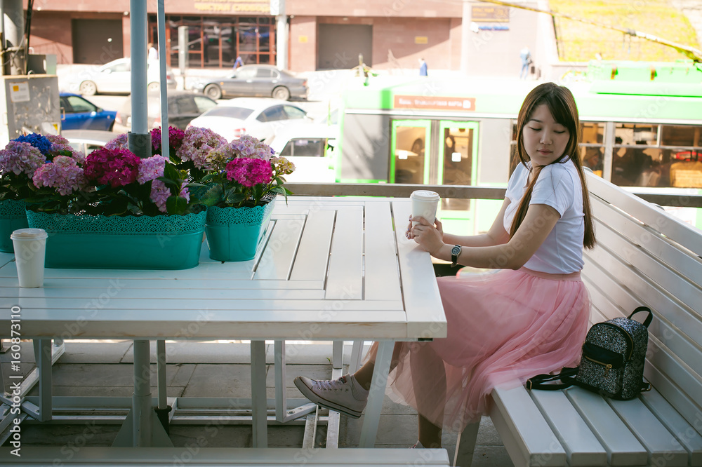 Cute asian young woman in summer cafe outdoors. girl In white T-shirt, with long hair in simple light cozy interior of restaurant Urban style, drinking coffee drink, sitting at table with flowers Cute asian young woman in summer cafe outdoors. girl In white T-shirt, with long hair in simple light cozy interior of restaurant Urban style, drinking coffee drink, sitting at table with flowers