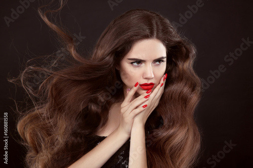 Beauty hair. Red manicure. Brunette girl with long shiny wavy hair. Beautiful model portrait with curly hairstyle and manicured nails isolated on studio dark background.