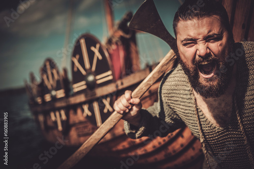 Viking with axe standing near drakkar on the seashore. photo