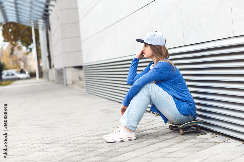 Young girl sitting on skateboard with mobile phone and headphone against modern gray wall.