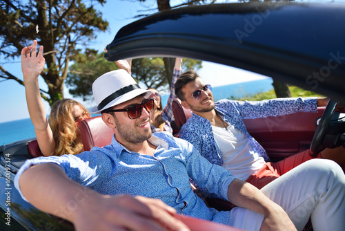 leisure, road trip, travel and people concept - happy friends driving in cabriolet car along country road