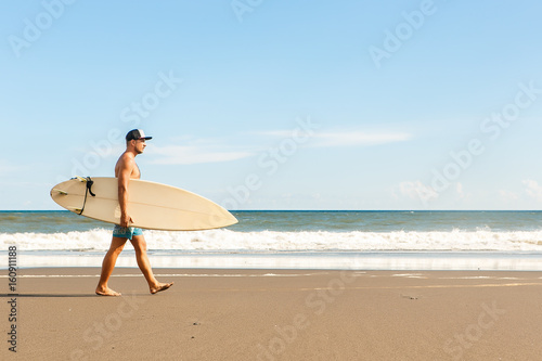 Handsome fit man walk with white blank surfboard wait for wave to surf spot at sea ocean beach. View from side. Concept of sport, fitness, freedom, happiness, new modern life, hipster, generation Y.