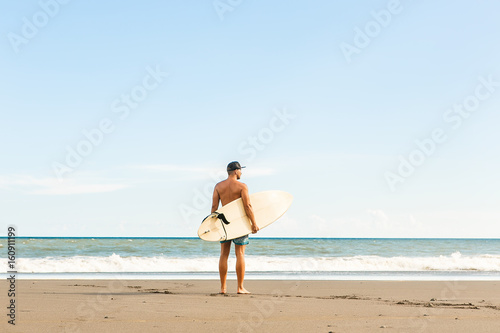 Handsome man stand with long surf surfing board wait on surf spot at sea ocean beach. View from back. White blank surfboard. Concept of sport, freedom, new modern life, generation Y.