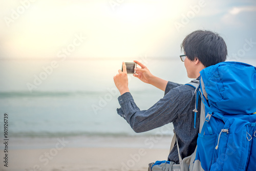 Young Asian backpacker man taking photos of beach and sea by smartphone, summer holiday vacation and travel tropical island concepts