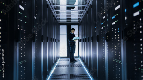 Male IT Engineer Works on a Laptop at the end of a Corridor in a Big Data Center. Rows of Rack Servers are Seen.
