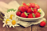 Sweet wild strawberries in plate with chamomiles on wooden background, toned