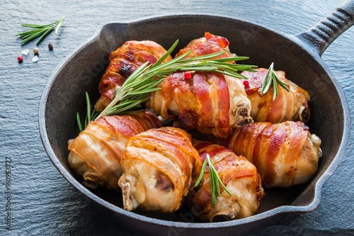 Bacon wrapped chicken drumsticks in a black cast-iron skillet on the stone background.