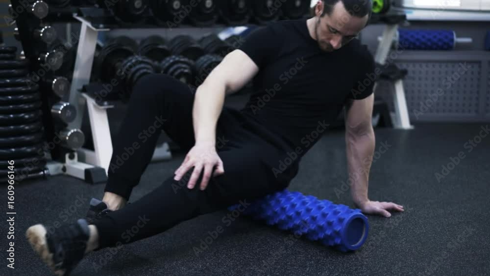 Close up of a man wearing black sportswear working with his buttock on a blue roll in a gym. Concept of fitness. Real time tilt down tilt up pan shot left to right close up shot Close up of a man wearing black sportswear working with his buttock on a blue roll in a gym. Concept of fitness. Real time tilt down tilt up pan shot left to right close up shot