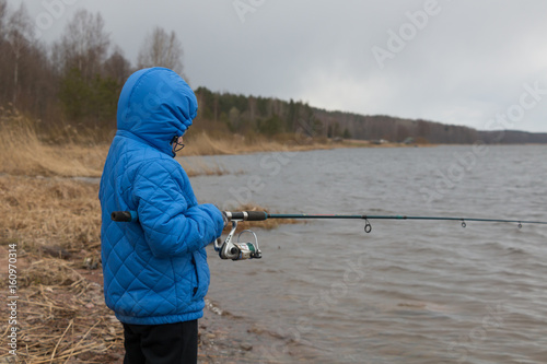 The boy in the blue jacket fishing on the lake. Cold weather.