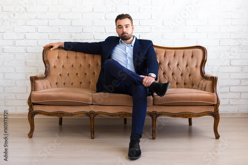 richness and success concept - handsome bearded man in business suit sitting on vintage sofa