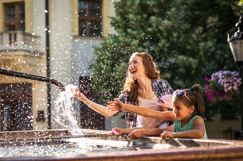 Mother and her daughter playing in the city square fountain. They sprayed with water. Refreshing on a hot summer day. 