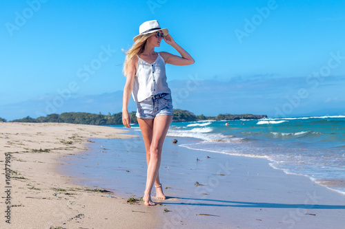 Young sexy woman with white hat walking on white sand beach a tropical Bali island at sunny day. Ocean cost.