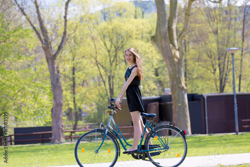 Portrait view of girl on bicycle wearing on black short dress. Young happy Woman riding along road on green spring outdoor Park. Sporty young girl riding a bicycle on a sunny morning