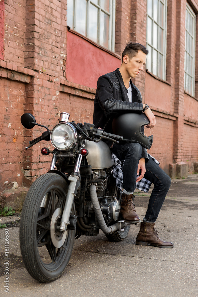 Handsome rider biker guy in black leather jacket, boots and style denim sit on classic style cafe racer motorcycle. Bike custom made in vintage garage. Brutal fun urban lifestyle. Outdoor portrait.