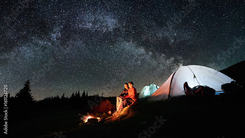 Night camping in the mountains. Man and woman tourists sitting at a campfire near two illuminated tents under beautiful night sky full of stars and milky way. Long exposure. Picture aspect ratio 16:9