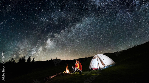 Night camping. Couple tourists sitting at a campfire near illuminated tent under incredible night sky full of stars and milky way. Long exposure. Picture aspect ratio 16:9