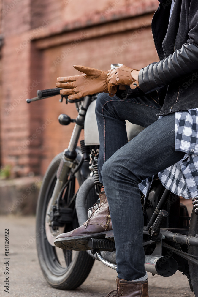 Obraz premium Zbliżenie na przystojny rowerzysta jeździec ręka facet stawia na skórzaną rękawiczkę, siedząc na motocyklu cafe racer w stylu klasycznym. Rower wykonany na zamówienie w garażu vintage. Brutalna zabawa w miejskim stylu życia. Portret na zewnątrz.