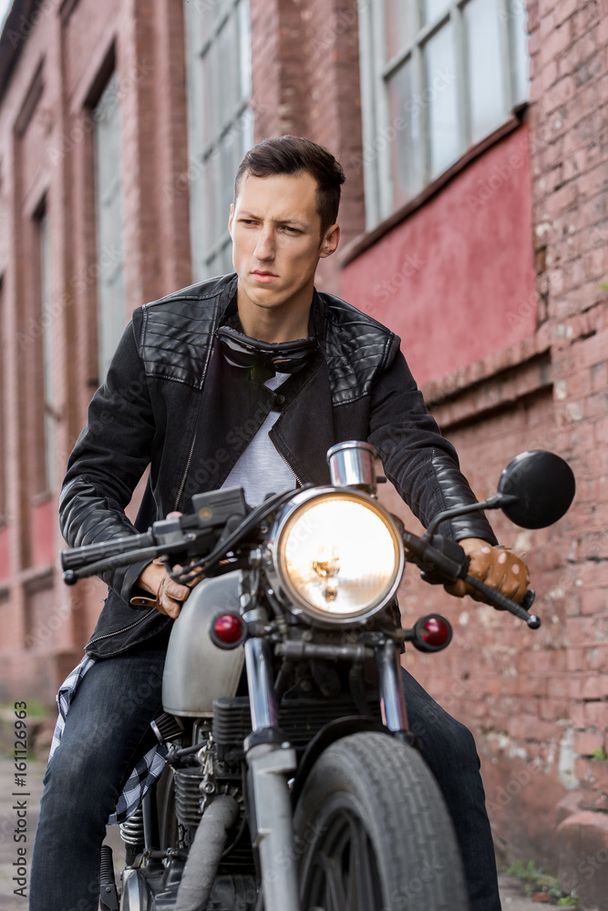 Handsome rider biker guy in black leather jacket, jeans, boots and gloves  sit on classic style cafe racer motorcycle. Bike custom made in vintage  garage. Brutal fun urban lifestyle. Outdoor portrait. Stock