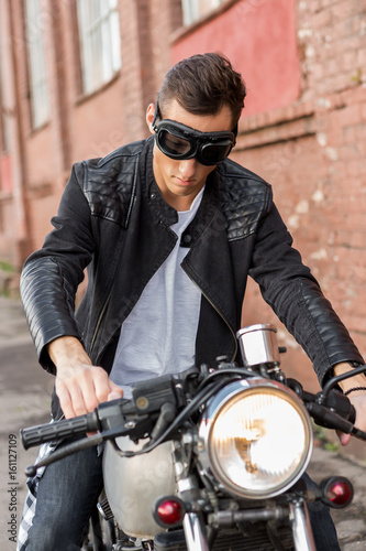 Handsome rider biker man in black leather jacket and protection googles sit on classic style cafe racer motorcycle. Bike custom made in vintage garage. Brutal fun urban lifestyle. Outdoor portrait.