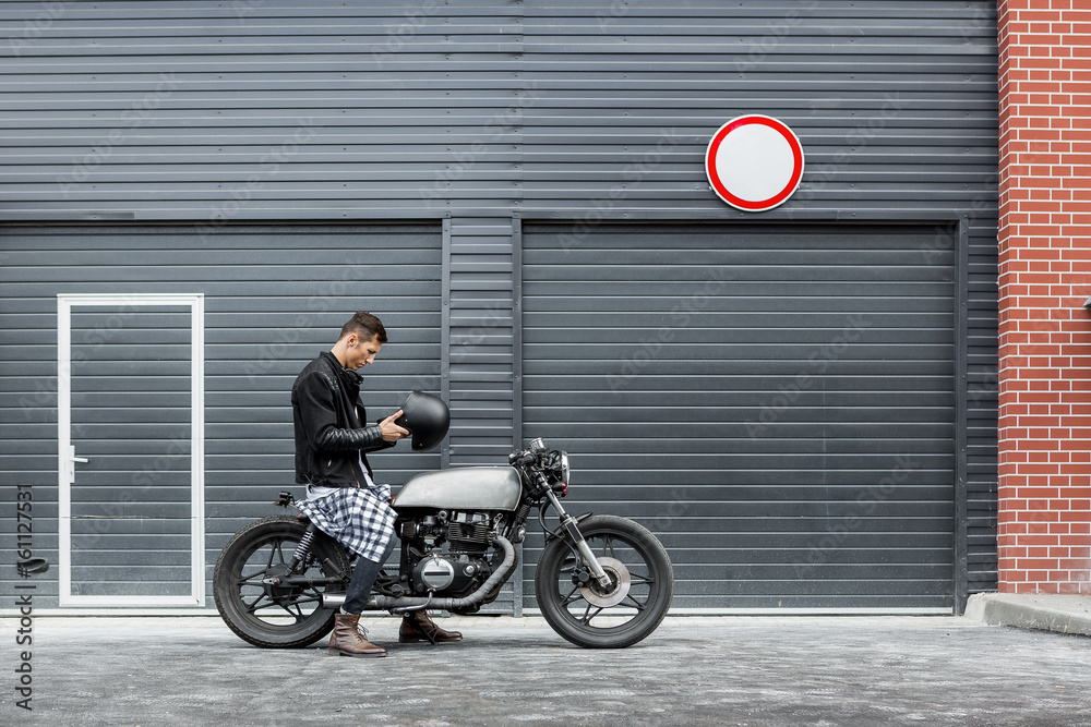 Fototapeta premium Handsome rider man take off black biker helmet while sit on classic style cafe racer motorcycle near industrial gates. Bike custom made in vintage garage. Brutal fun urban lifestyle. Outdoor portrait.