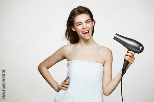 Cheerful beautiful girl in towel smiling laughing singing with hair dryer making funny face over white background. Beauty spa and cosmetology.
