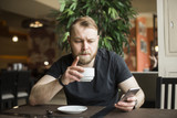 Young bearded businessman,dressed in gray cardigan,sitting at table in cafe and use smartphone. Man holding smartphone and looking at its screen. Man using gadget. Guy browsing internet on smartphone