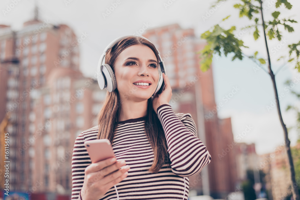 Carefree young lady is listening to favourite song on her smartphone outdoors, walking on the spring street, wearing cozy outfit, modern headphones