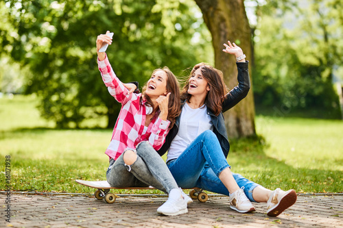 Cheerful young women siting on longboard in park posing for selfie photo