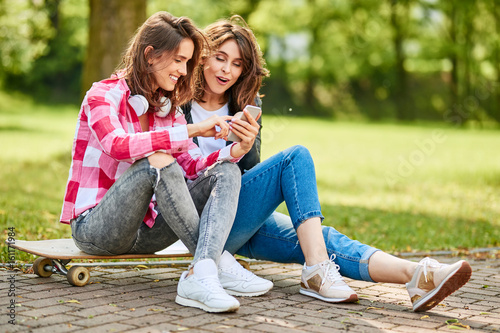Joyful female friends sitting on longboard looking at smart phone