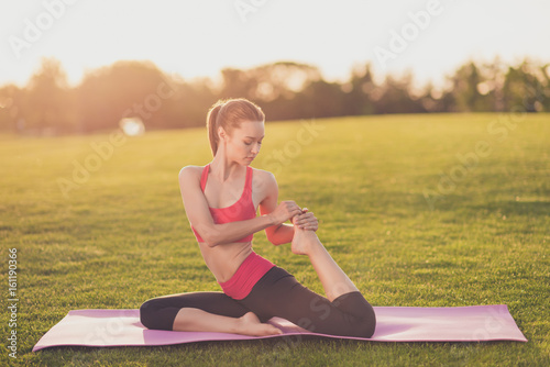 Young yoga trainer is making stretching sitting outdoors in spring park on a purple mat on nice green grass, so relaxed and healthy, wearing modern pink and black sport outfit, with tail