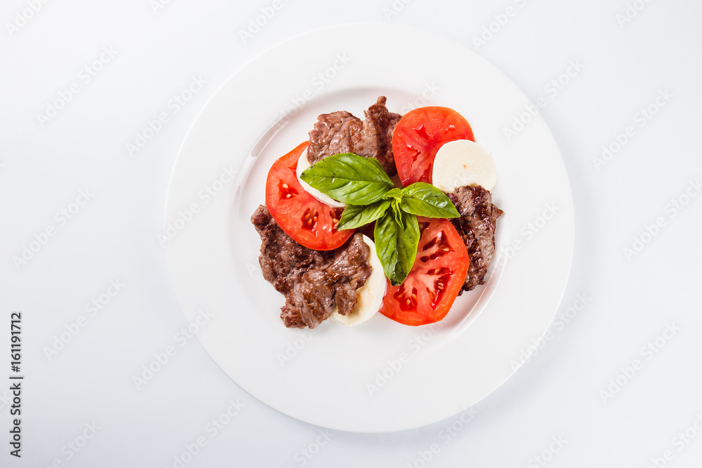Meat salad. Fried beef with mozzarella, tomato and basil on a white plate on a light background (top view close) Meat salad. Fried beef with mozzarella, tomato and basil on a white plate on a light background (top view close)