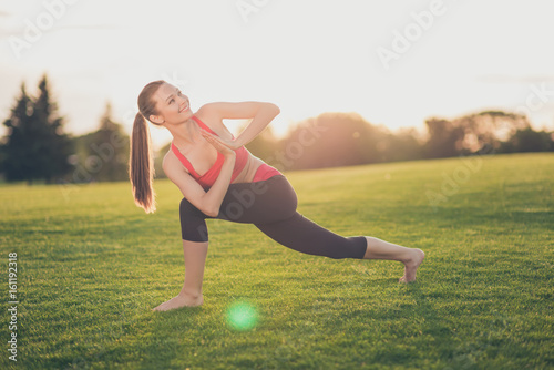 Namaste! Young cheerful woman is doing complex yoga exercise outdoors in a spring morning park on a nice green grass, in trendy sport wear, with tail