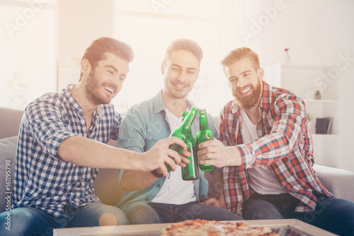 Cheers men! Bachelor men`s life. Close up of three happy friends, clinking their beers, smiling, alll are in casual outfits. They are fans of sports games as football, basketball, hockey, baseball photo