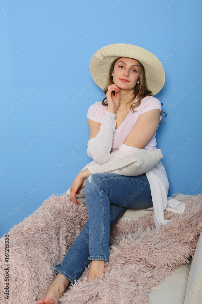 Feminine woman sitting on beige chair, boudoir, in straw hat on blue background