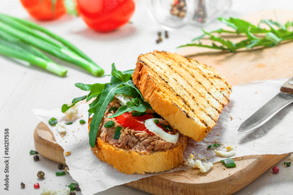 Sandwich made of grilled toasts, tuna salad, tomato, onion and arugula on a white wooden table. Delicious healthy meal with ingredients. Sandwich made of grilled toasts, tuna salad, tomato, onion and arugula on a white wooden table. Delicious healthy meal with ingredients.