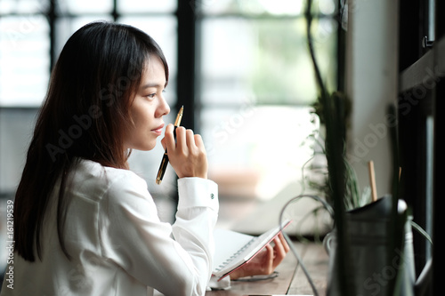 young women holding pen for writing on notebook. her sitting by the window at library room in morning. image for education,business,people,interior and portrait concept photo