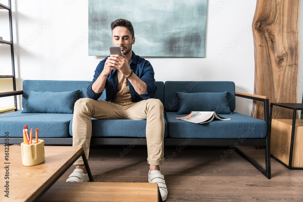 Obraz premium Concentrated youthful bearded guy using his smart phone in apartment