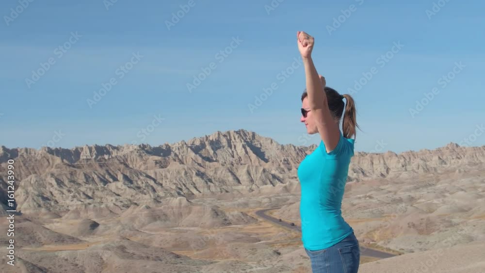 SLOW MOTION: Female tourist hiking Badlands National park and victoriously raising her arms up, overlooking pointy sandstone mountains on sunny day. Cheerful woman celebrating reaching mountaintop SLOW MOTION: Female tourist hiking Badlands National park and victoriously raising her arms up, overlooking pointy sandstone mountains on sunny day. Cheerful woman celebrating reaching mountaintop