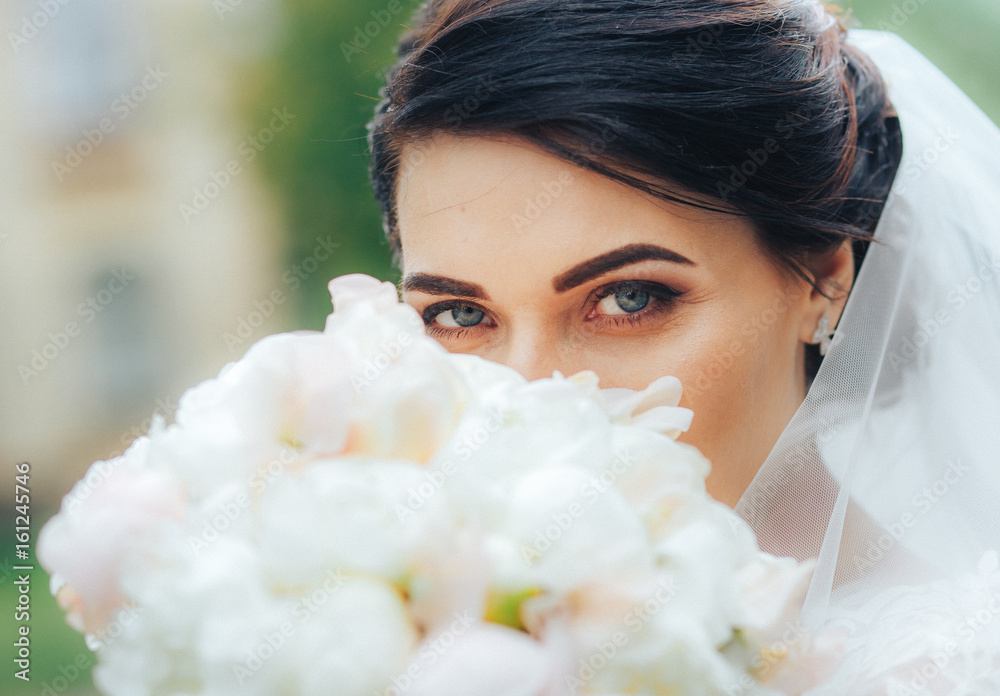 Naklejka premium A beautiful warrior bride with blue eyes looks at the camera hiding behind a bouquet of white flowers