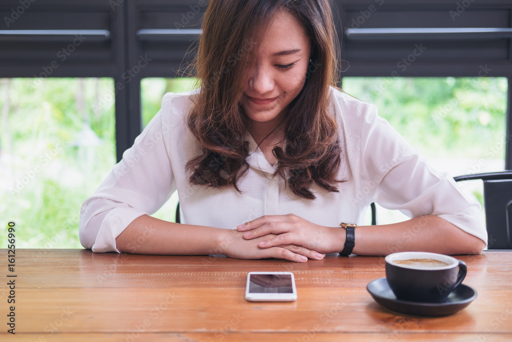 A beautiful Asian woman with smiley face looking at smart phone on wooden table in modern cafe with green nature background A beautiful Asian woman with smiley face looking at smart phone on wooden table in modern cafe with green nature background