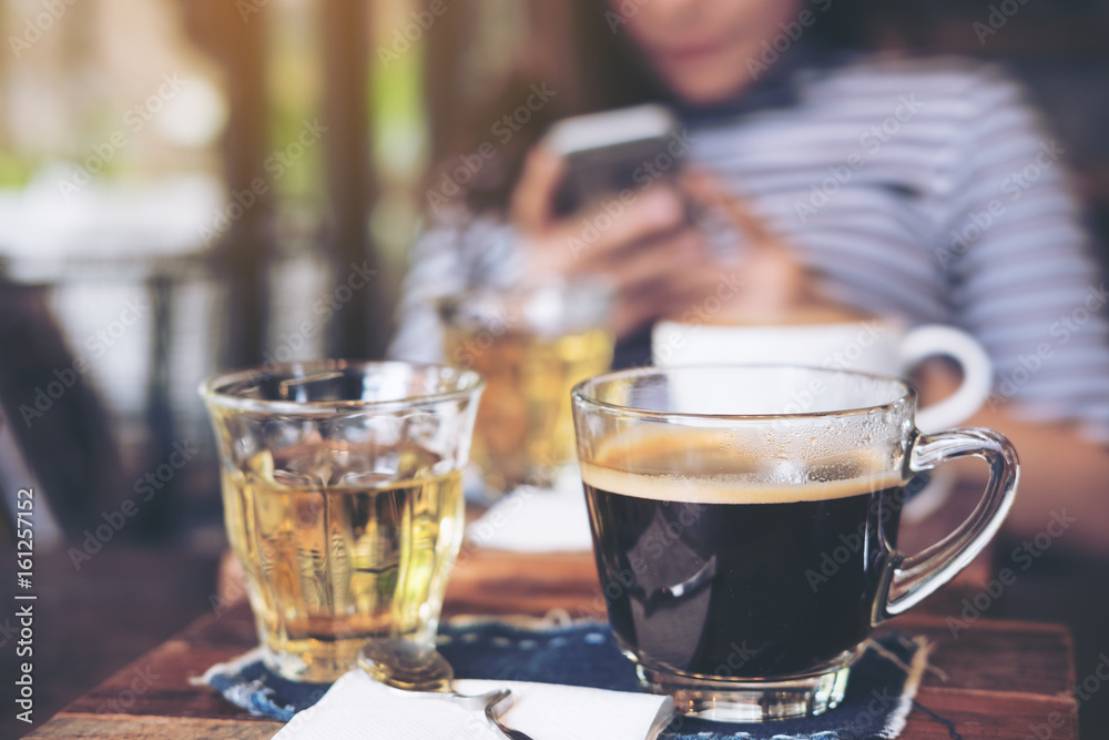 Closeup image of cups of hot coffee and tea on vintage wooden table in cafe with blur woman using smart phone in background Closeup image of cups of hot coffee and tea on vintage wooden table in cafe with blur woman using smart phone in background