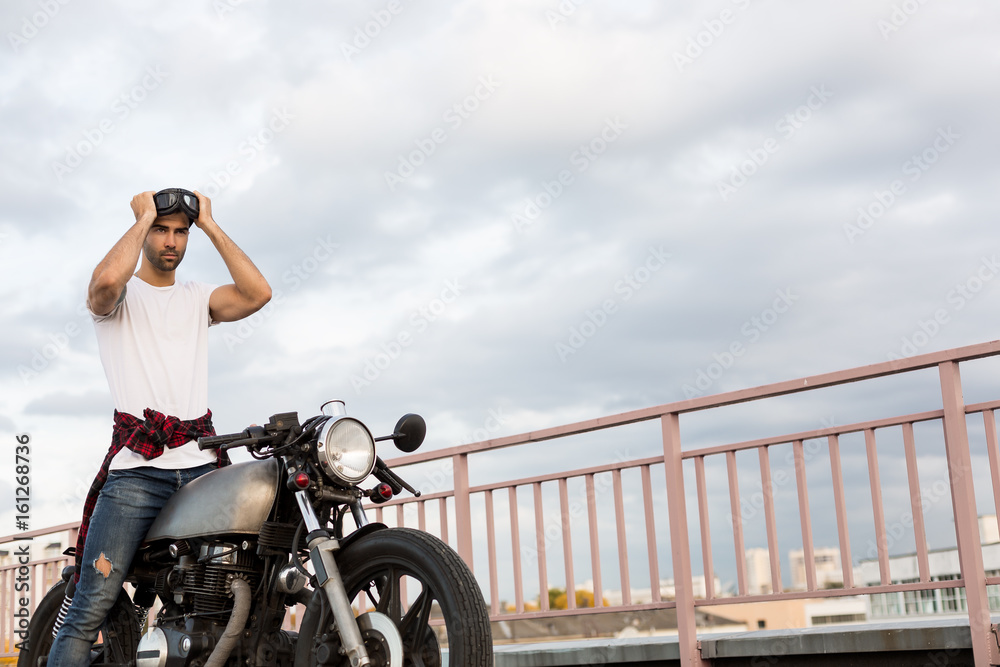 Handsome rider man with beard and mustache posing while put on black moto  sunglasses near classic style biker cafe racer motorcycle. Bike custom made  in vintage garage. Brutal fun urban lifestyle. Stock