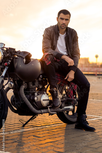 Handsome rider guy with beard and mustache in black leather biker jacket, boot and jeans sit on classic style cafe racer motorcycle. Bike custom made in vintage garage. Brutal fun urban lifestyle.