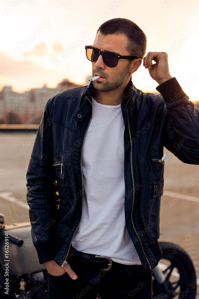 Handsome rider man with beard and mustache in black biker jacket, white t- shirt and sunglasses smoking cigaret near classic style cafe racer  motorbike on rooftop at sunset. Brutal fun urban lifestyle. Stock