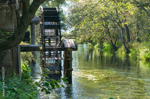 Fotografija Water wheel mill on river with green trees on the background